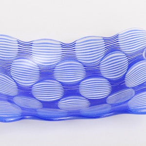 Puede incluir: Un plato de vidrio azul y blanco con forma rectangular ondulada. El plato presenta un patrón de rayas blancas horizontales sobre un fondo azul, creando un efecto texturizado similar a burbujas. El plato probablemente se utiliza con fines decorativos.