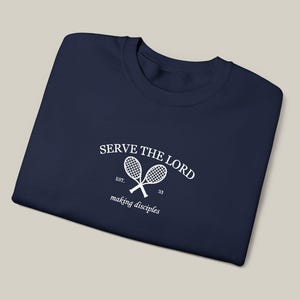 Könnte beinhalten: Marineblaues T-Shirt mit dem weißen Schriftzug "SERVE THE LORD" und zwei gekreuzten Tennisschlägern. Die Worte "making disciples" und die Zahl "33" sind ebenfalls weiß gedruckt.