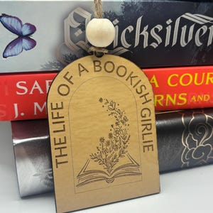 May include: A wooden bookmark with the text "THE LIFE OF A BOOKISH GIRLIE" and an illustration of an open book with floral accents. The bookmark is hanging in front of a stack of books.