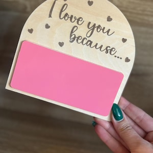 May include: Wooden sign with the text "I love you because..." and small heart accents. A bright pink rectangular insert is set into the wooden frame. The sign is held by a hand with green nail polish.