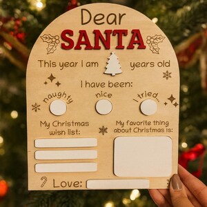May include: Wooden Christmas sign with the words "Dear Santa" in red. It includes spaces to write age, behavior, wish list, and favorite things about Christmas. The sign is decorated with snowflakes and a Christmas tree.