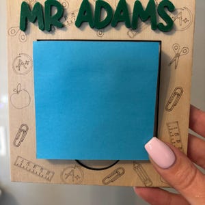 Puede incluir: Un marco de madera con una nota adhesiva azul y el nombre "MR ADAMS" en letras verdes. El marco está decorado con dibujos de temática escolar, incluyendo lápices, reglas, tijeras y manzanas.