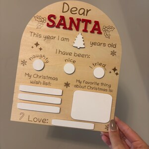 May include: Wooden Christmas sign with the words "Dear Santa" in red. The sign includes spaces to write age, wish list, and favorite things about Christmas. Features a Christmas tree and snowflake accents.