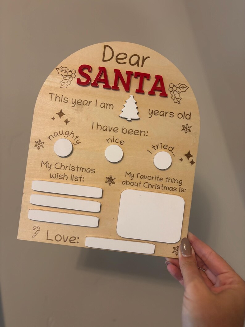 May include: Wooden Christmas sign with the words "Dear Santa" in red. The sign includes spaces to write age, wish list, and favorite things about Christmas. Features a Christmas tree and snowflake accents.