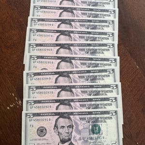 May include: A stack of ten United States five-dollar bills arranged in numerical order, featuring the portrait of Abraham Lincoln, the text "The United States of America," and serial numbers ranging from QF 45803291 B to QF 45803300 B.