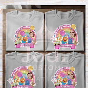 May include: Four gray t-shirts with a colorful graphic design. Each shirt features a cartoon image of a girl, a rainbow, and the text "Hi Friends! Can You Say". The shirts are labeled "Brother", "Grandma", "Grandpa", and "Ahmiya".
