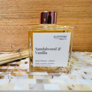 Puede incluir: Una botella de vidrio transparente con una tapa dorada llena de un líquido marrón claro. La botella está etiquetada con "summer candle co" y "Sandalwood & Vanilla Reed Diffuser | 200ml". La botella está sobre una superficie de azulejos blancos y marrones.