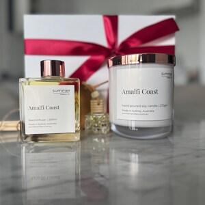 May include: A gift set featuring a reed diffuser and a candle, both labeled "Amalfi Coast." The candle is in a clear glass jar with a rose gold lid. The diffuser is in a square glass bottle with a rose gold cap. A white gift box with a red ribbon is in the background.