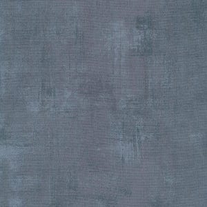 May include: A close-up of a textured fabric with a mottled appearance. The fabric is primarily a muted gray-blue color, with lighter and darker areas creating a distressed, painterly effect. The surface has a subtle, woven texture.