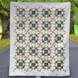 Op de afbeelding: Een wit quilt met een geometrisch patroon van groene, oranje en zwarte sterren. Het quilt hangt aan een waslijn voor een grasveld met bomen op de achtergrond.