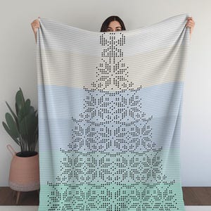 May include: A large, rectangular blanket with a pixelated Christmas tree design. The blanket features a gradient of colours, transitioning from light blue to teal, with a white and black tree pattern. The blanket is held up by a person.