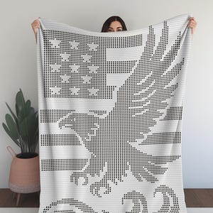 May include: A white blanket with a black graphic design featuring an eagle, stars, and stripes. The blanket has a pixelated appearance, with the design created using small black squares. The overall theme is patriotic.