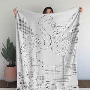 May include: A white blanket with a black dotted design featuring two flamingos, flowers, and foliage. The blanket is held up, showcasing the intricate pattern. The design is reminiscent of cross-stitch or pixel art.