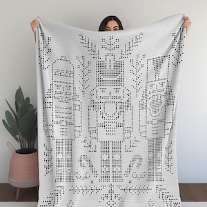 May include: A white blanket with a black geometric pattern. The design features stylized figures, trees, and snowflake-like shapes. The blanket is held up, showcasing the intricate details of the woven design.