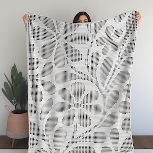 May include: A white blanket with a black floral pattern. The design features large stylized flowers and leaves, created with a pixelated effect. The blanket is held up, showcasing the full pattern.