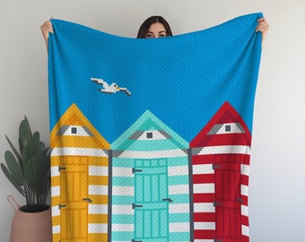 May include: A beach-themed crochet blanket featuring three colorful beach huts with white stripes, a blue sky, and a white seagull flying overhead. The blanket has a sandy beach scene at the bottom with white waves.