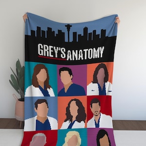Op de afbeelding: Een deken met een pixelontwerp met de tekst "GREY'S ANATOMY" boven een raster van karakterportretten. De deken heeft een blauw skyline-ontwerp aan de bovenkant.