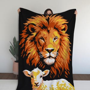 May include: A black blanket featuring a pixelated design of a lion's head in shades of orange and yellow above a lamb in white and yellow. The blanket is held up against a white wall, showcasing the detailed animal imagery.
