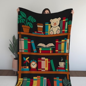 May include: Black throw blanket with a pixelated bookshelf design. The blanket depicts colourful books, potted plants, a teddy bear, a coffee cup, and an alarm clock. The bookshelf is brown, and the books are various colours.
