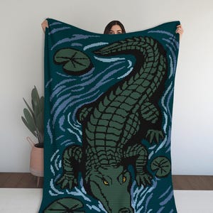 May include: A dark teal blanket with a pixelated design of a green and black alligator, with yellow eyes. The alligator is set against a water background with lily pads. A cosy home decor item.
