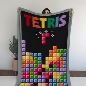 May include: A Tetris-themed blanket with a black backdrop, displaying the game's logo in rainbow hues. The design features pixelated falling blocks in multiple colours. The blanket has a grey border.
