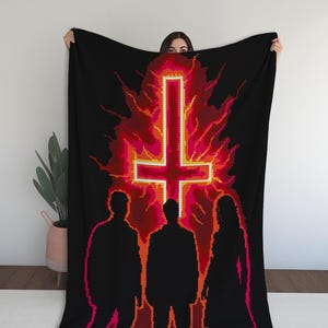 May include: Black blanket featuring a pixelated design of an inverted cross in a fiery, red and orange background. Silhouettes of three figures stand below the cross. The blanket is held up by a person.