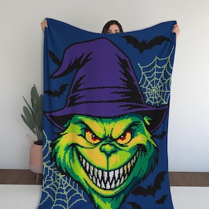 May include: A blue blanket featuring a pixelated image of a green creature with a purple witch hat, a wide grin, and red eyes. Black bats and a spiderweb design are in the background. The blanket is held up by a person.