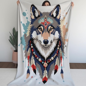Peut inclure: Une couverture tissée blanche présentant une grande tête de loup de style pixel art au centre. Le loup est orné de motifs géométriques colorés sur le front et d'un collier de perles et de plumes dans des tons de rouge, bleu et beige sur fond blanc.