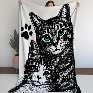 Peut inclure: Un plaid blanc orné d'un grand motif pixelisé en noir et blanc représentant deux chats aux yeux bleu sarcelle. Le design comprend une petite empreinte de patte noire à gauche de la tête du plus grand chat, dans un style en blocs distinct.