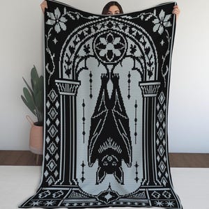 May include: A rectangular throw blanket featuring a black and light gray pixelated design. The pattern displays a bat hanging upside down within a central arched architectural frame, flanked by decorative geometric borders and floral motifs.