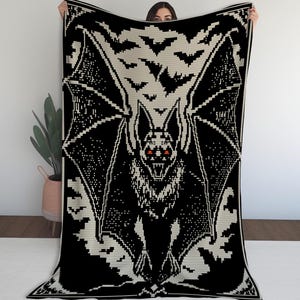 May include: Black and cream-colored crochet blanket featuring a large bat with outstretched wings and red eyes. The bat is surrounded by smaller bat silhouettes. The blanket is a cozy, decorative item.