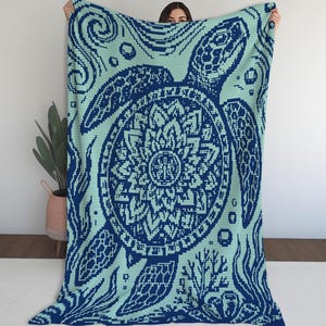Puede incluir: Una manta rectangular grande con un diseño de arte de píxeles de una tortuga marina con un patrón de caparazón estilo mandala. La manta es de color verde menta claro y azul marino oscuro, representando a la tortuga rodeada de coral y burbujas.