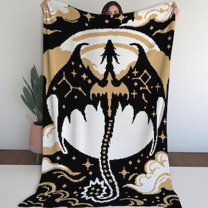 Könnte beinhalten: Eine schwarz-weiß-goldene Häkeldecke mit einem Drachendesign. Der Drache hat ausgebreitete Flügel und einen langen Schwanz, vor dem Hintergrund eines Mondes und eines Sternenhimmels. Die Decke wird von einer Person gehalten.
