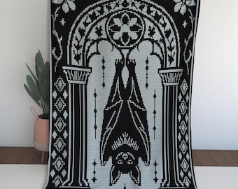 May include: A rectangular throw blanket featuring a black and light gray pixelated design. The pattern displays a bat hanging upside down within a central arched architectural frame, flanked by decorative geometric borders and floral motifs.