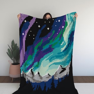 Peut inclure: Une couverture en crochet noire et bleue avec un motif pixélisé des aurores boréales au-dessus d'une chaîne de montagnes.