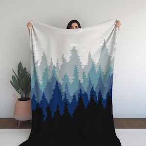 Peut inclure: Une couverture avec un motif de forêt pixélisée. Le motif passe du blanc à des nuances de bleu, pour finir par une base noire. La couverture est tenue, montrant le motif et la texture complets.