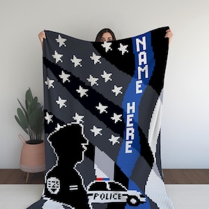 May include: A blue and white crochet blanket featuring a thin blue line, an American flag, and a silhouette of a police officer standing next to a police car. The text "NAME HERE" is written vertically on the blue line. The text "POLICE" is written on the police car.
