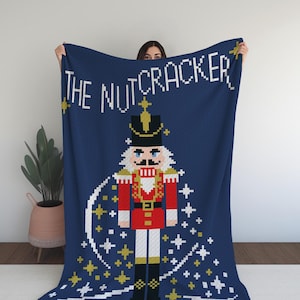 以下が含まれることがあります： 剣を持ったくるみ割り人形のピクセル画像が描かれた青いニットの毛布。毛布の上部には白い文字で「The Nutcracker」と書かれています。