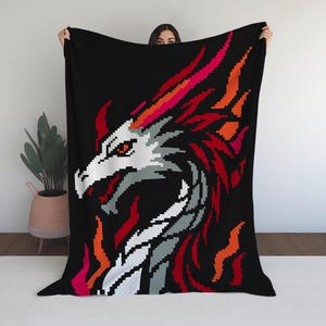 May include: Black blanket featuring a pixelated dragon design. The dragon has a white head, gray accents, and red and orange flames. The blanket is held up against a white wall, showcasing the design.