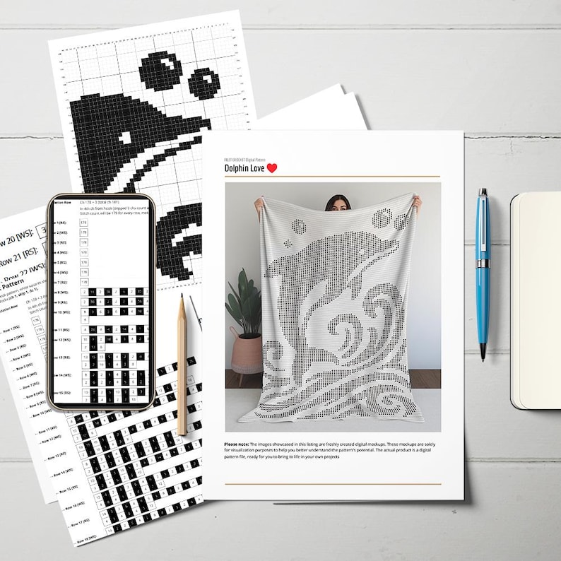 May include: A white blanket with a black dolphin design, held up in front of a person. The design includes bubbles and waves. Accompanying the blanket are printed crochet patterns, a smartphone displaying a pattern, a pencil, a pen, and a notebook.