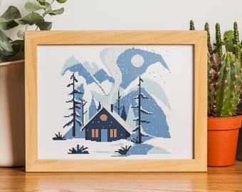 May include: A cross-stitch pattern featuring a blue and white winter scene with a small cabin, trees, and mountains. The cabin has a brown door and windows. The sky is a light blue with white snowflakes.
