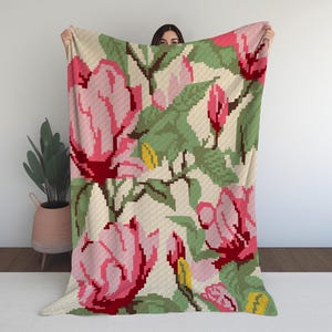 May include: A large, pixelated floral blanket with a repeating pattern of pink roses, green leaves, and yellow accents on a cream background. The blanket is held up, showcasing the design. The blanket is a cozy home decor item.