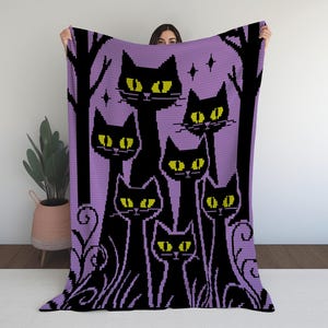 Puede incluir: Una manta morada con un diseño de gatos negros con ojos amarillos. Los gatos están colocados sobre un fondo de árboles y estrellas estilizados. La manta está sujeta por una persona. Un artículo acogedor.