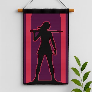 Puede incluir: Un tapiz de pared pixelado negro que presenta la silueta de una persona sosteniendo un objeto largo sobre sus hombros. La figura se alza sobre un fondo de rayas verticales rojas y moradas. El tapiz está montado en una barra de madera.