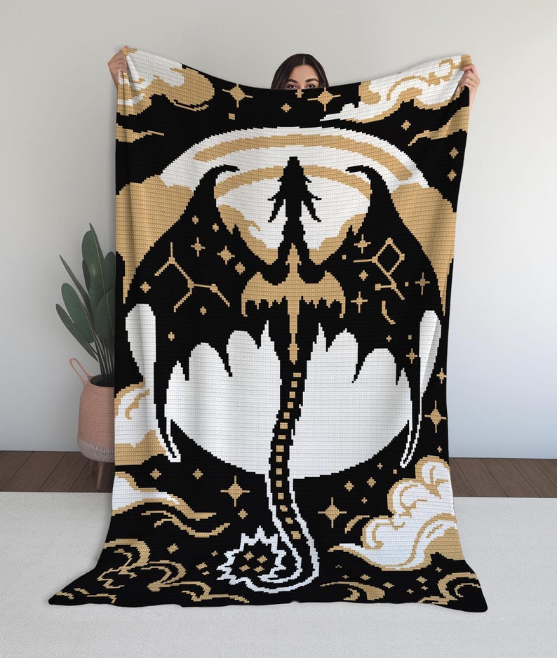 May include: A black, white, and gold crochet blanket featuring a dragon design. The dragon has outstretched wings and a long tail, set against a moon and starry sky. The blanket is held up by a person.
