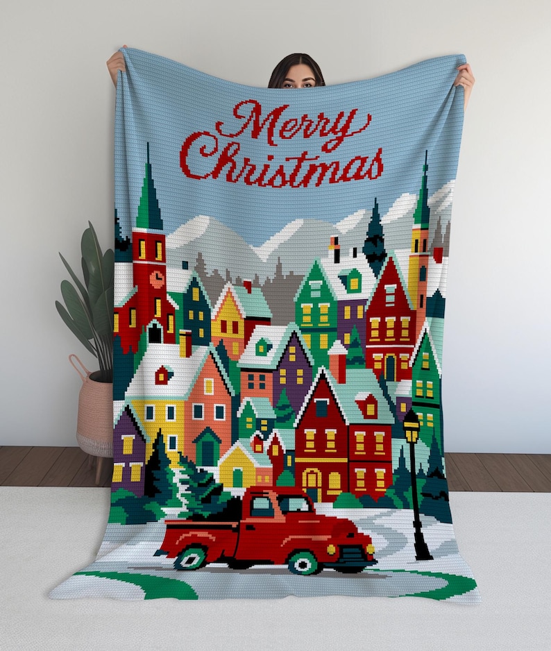 May include: A blue blanket with a Christmas village scene. The blanket features a red truck carrying a Christmas tree, colorful houses, and the words "Merry Christmas" in red script. The background includes snow-covered mountains and a light blue sky.