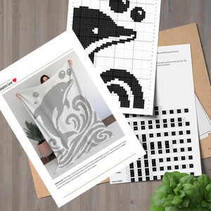 May include: A white blanket with a black dolphin design, held up in front of a person. Accompanying the blanket are several sheets of paper with a black and white dolphin pixel art pattern and a cross-stitch pattern. The blanket has the words "Dolphin Love" on it.