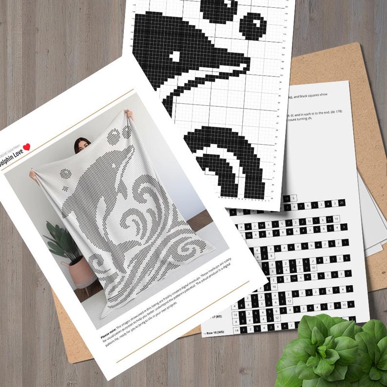 May include: A white blanket with a black dolphin design, held up in front of a person. Accompanying the blanket are several sheets of paper with a black and white dolphin pixel art pattern and a cross-stitch pattern. The blanket has the words "Dolphin Love" on it.