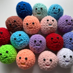 May include: A collection of 14 crocheted plush balls in various colors, each with a smiling face and black eyes. The balls are arranged in a circle on a white background.