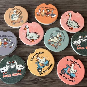 May include: A collection of ten car coasters with various goose-themed designs. The coasters feature phrases like "Just a Silly Goose" and "Goose Bumps," along with illustrations of geese on scooters and in various outfits. The coasters are round with a small notch.